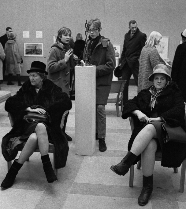 Svartvitt, äldre fotografi av två damer i hattar som sitter i varsin stol. Bakom damerna beskådar människor olika föremål i en utställningshall.