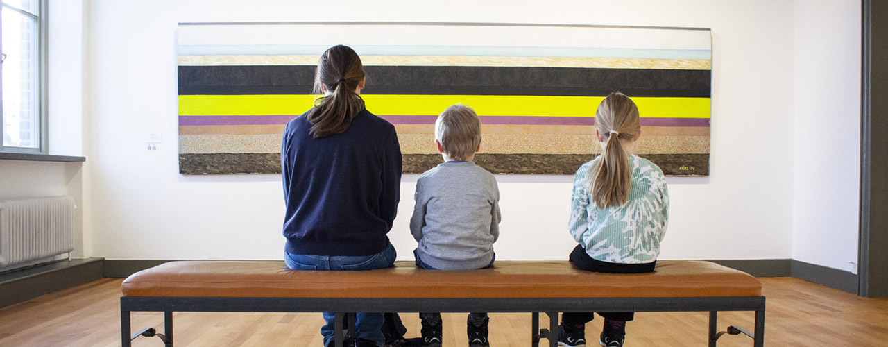 Besökare på Hallands Konstmuseum sitter på bänk och tittar på en målningen Halländskt landskap av Olle Kåks.
