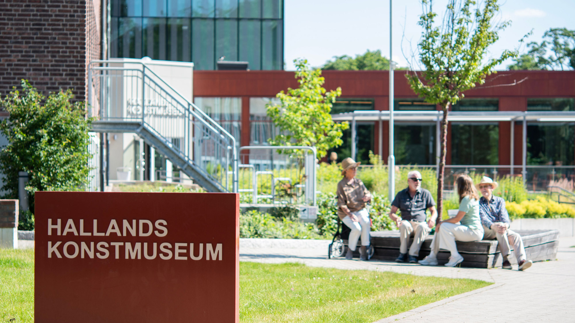 Skylt med texten Hallands Konstmuseum i museets trädgård. Besökare sitter i solen i bakgrunden.