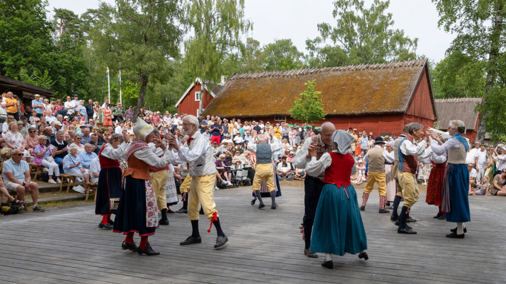 Folkdräktsklädda par från dansar inför en stor publik på Hallandsgårdens midsommarfirande.