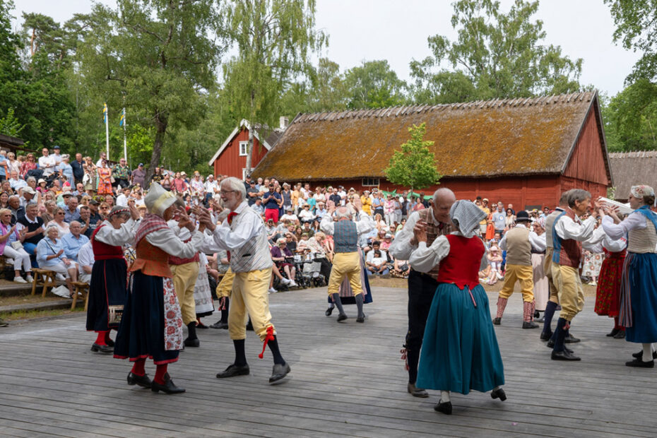 Folkdräktsklädda par från dansar inför en stor publik på Hallandsgårdens midsommarfirande.