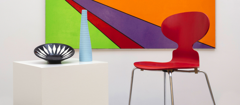 Stilleben med grafisk målning i bakgrunden bestående av triangulära fält i skarpa färger. I förgrunden står designföremål i form av en stol, vas och skål.