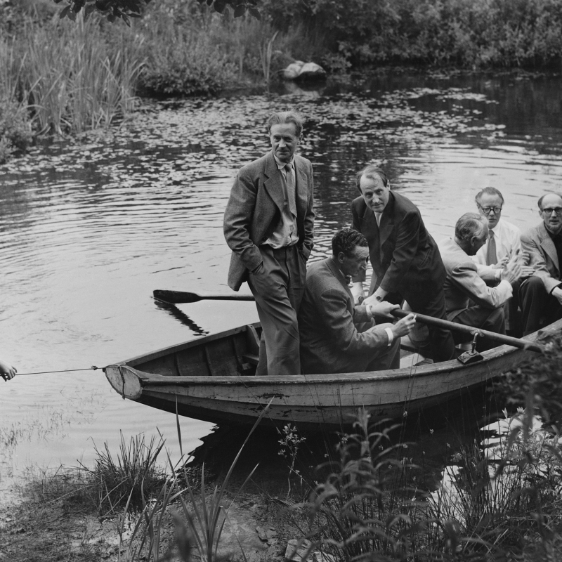Svartvitt, äldre fotografi med sex män i en eka. En pojke står vid vattenbrynet och drar iland ekan.